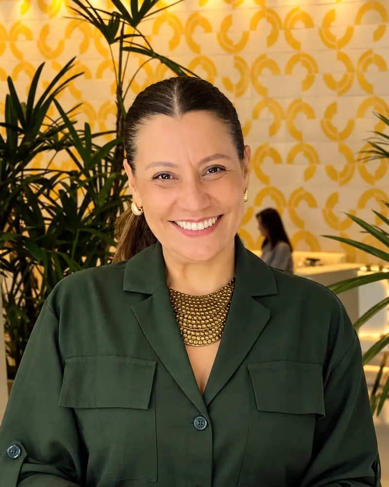 Profª. Érika Gadelha sorrindo, de camisa verde escura, em frente a uma parede com grafismos amarelos e plantas