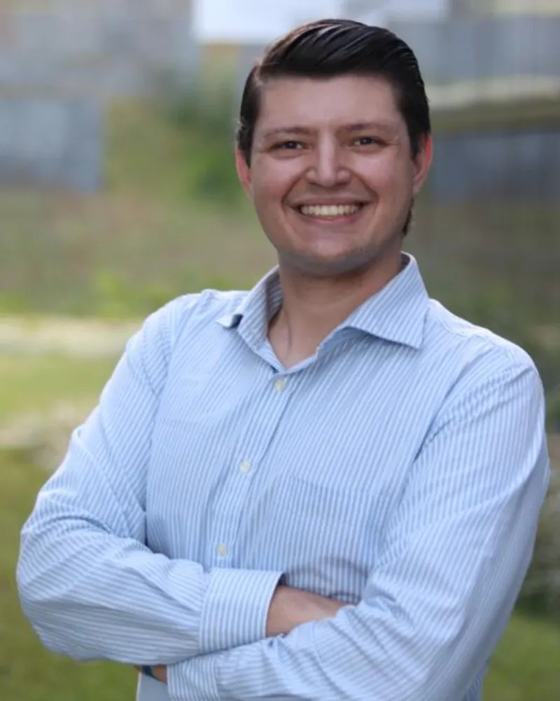 Prof. Dr. Marcos Costa sorrindo, de braços cruzados e camisa azul clara, em área externa